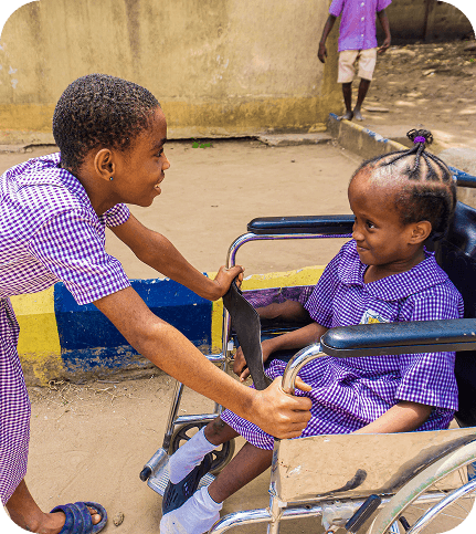 Girl smiles at classmate in a wheelchair outdoors.